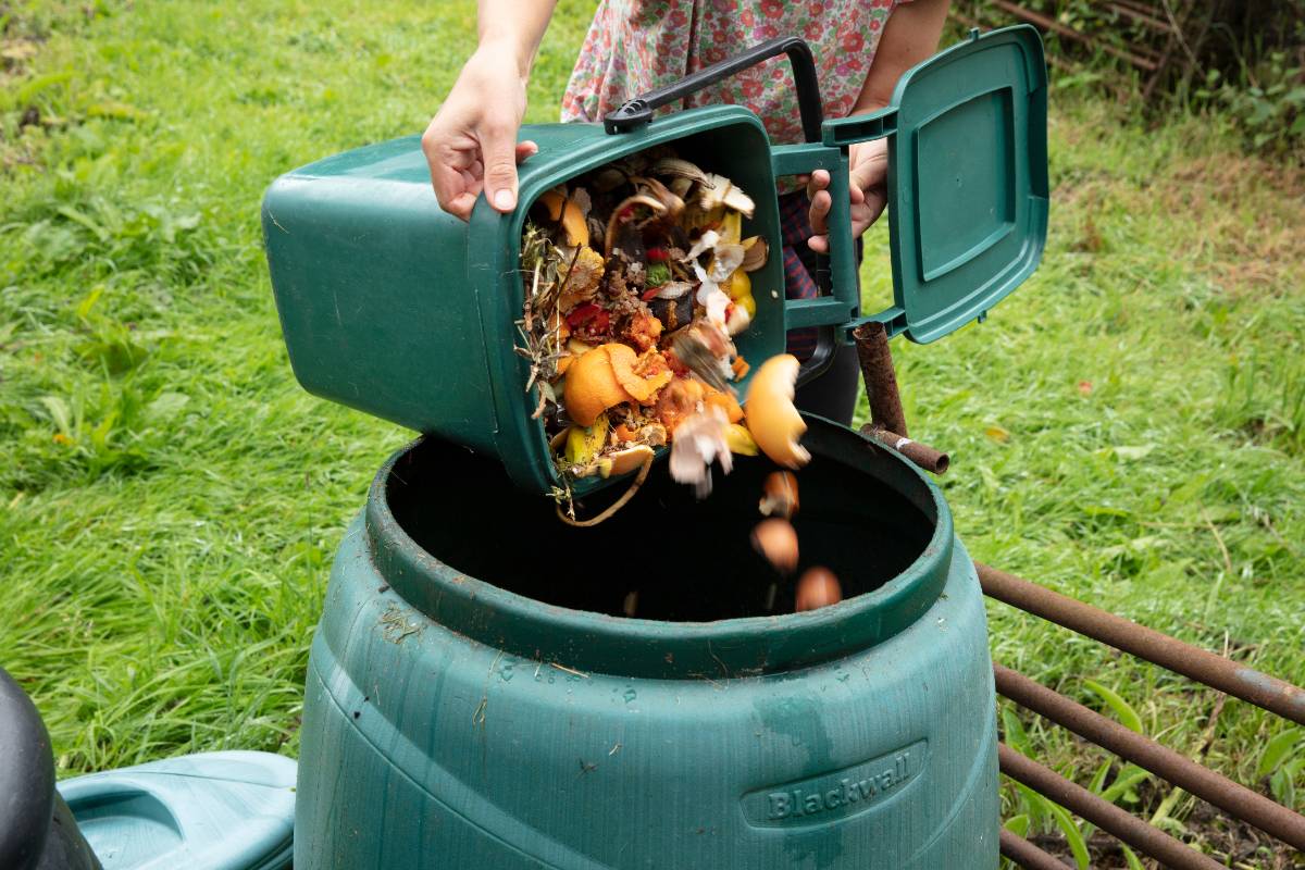 Person empties kitchen scraps from a compost caddy into an outdoor compost bin, showing fruit peels and food waste.