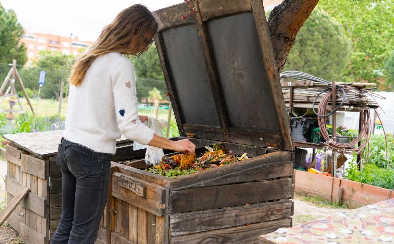 Woman adds kitchen scraps to a wooden backyard compost bin in a garden, demonstrating home composting and organic waste recycling.