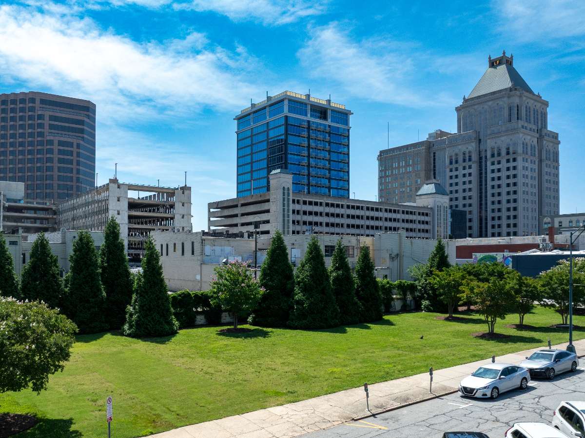 Spring midday view of downtown Greensboro skyline with office towers, parking garage, green lawn, trees, and parked street cars below.