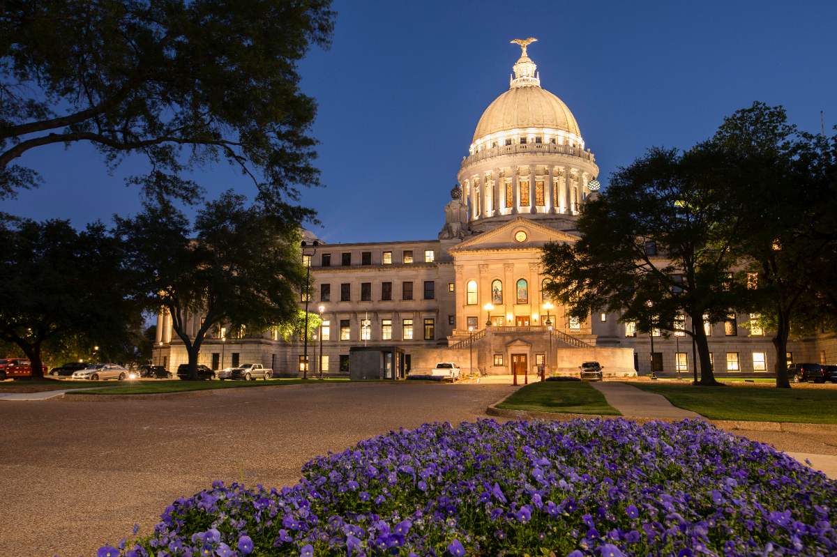 Spring twilight at Jackson Mississippi State Capitol, blooming purple flowers blanket foreground, trees budding softly frame illuminated domed building tonight.