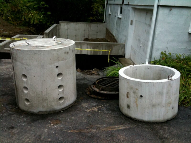 Precast concrete dry well components placed near a house foundation, ready for installation to manage yard drainage and prevent water pooling.