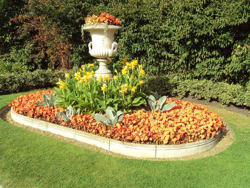 A neatly manicured flower bed with vibrant orange and yellow blooms arranged around a central decorative white urn planter. The garden bed is bordered by stone edging and surrounded by lush green grass and trimmed hedges.