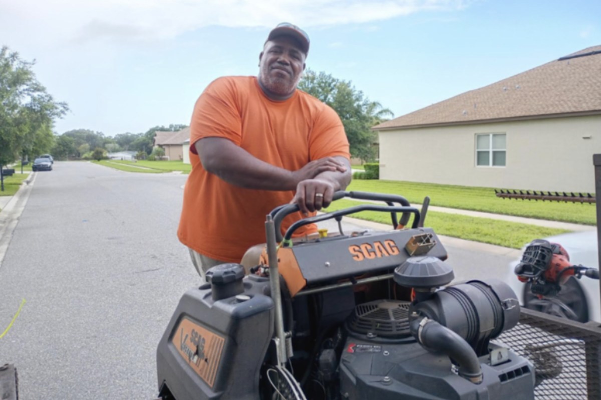 A man wearing an orange T-shirt and a cap stands outdoors on a residential street, leaning on a SCAG lawnmower. The background includes a sidewalk, green lawns, trees, and houses under a blue sky with light clouds. A leaf blower is visible on a trailer behind the lawnmower.