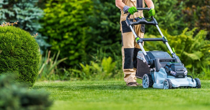 A person wearing work gloves and overalls is mowing a well-kept lawn with a modern electric lawn mower