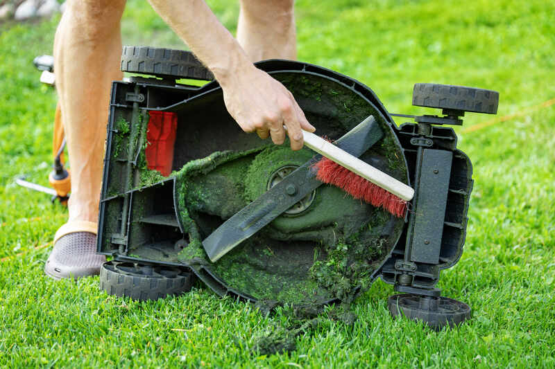 A person is cleaning the underside of a lawn mower blade with a brush on a grassy lawn