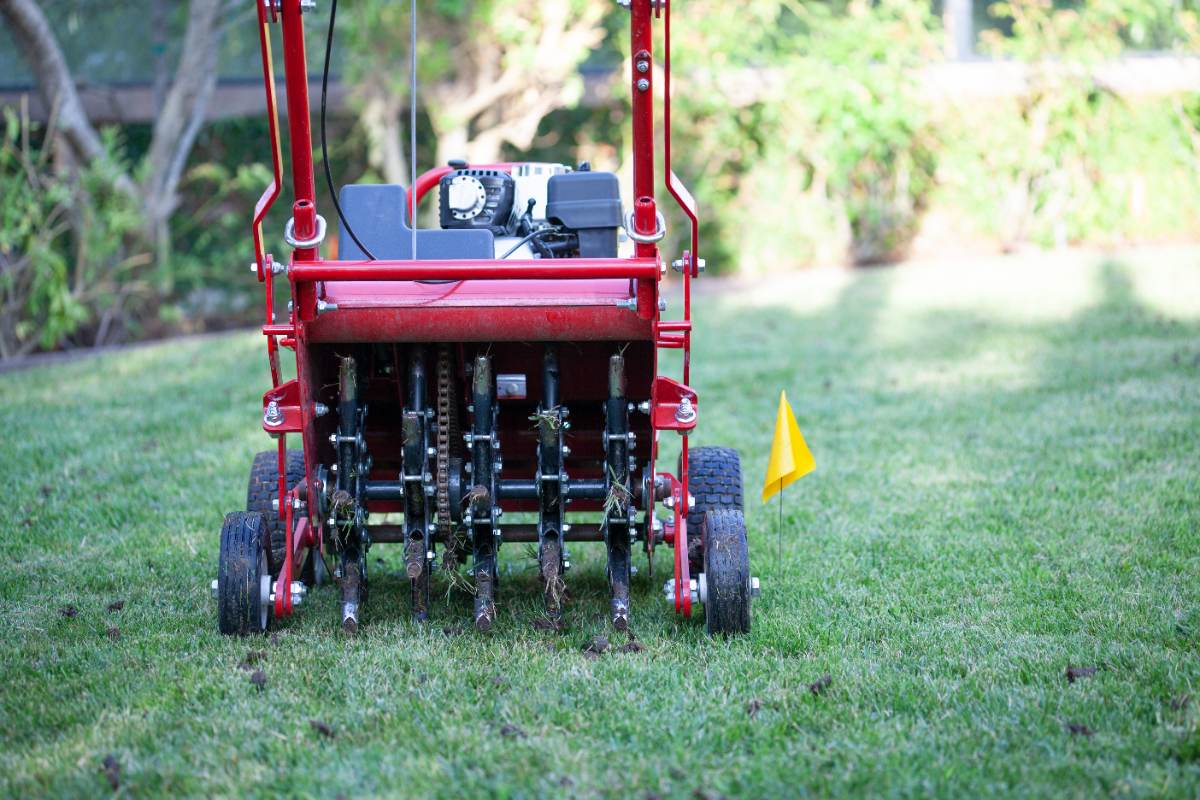 Rear view of a core lawn aerator machine in use on a grassy lawn, with soil plugs visible and a yellow marker flag nearby.