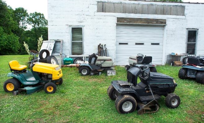 Numerous old lawn mowers sit on a lawn in front of a white building for sale.