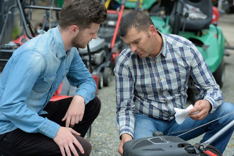Two men inspecting a lawn mower in an outdoor store setting, discussing its condition before a trade.