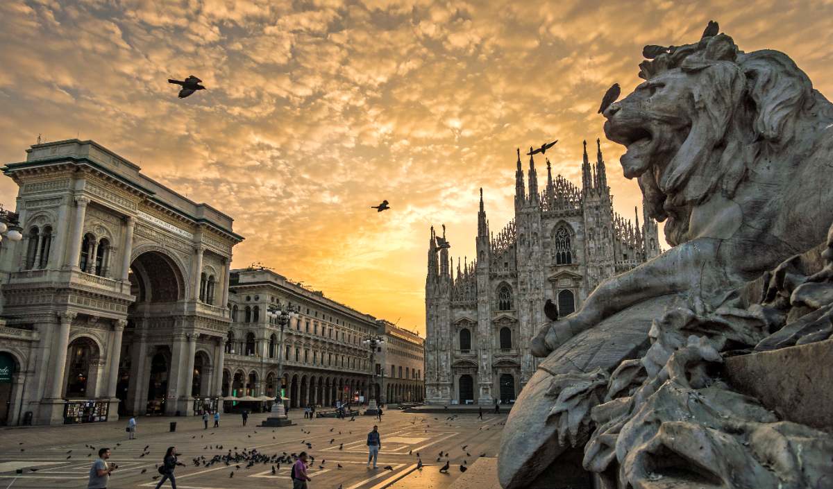 A lion sculpture sits in front of Milan Cathedral, an example of gothic architecture, at sunset.