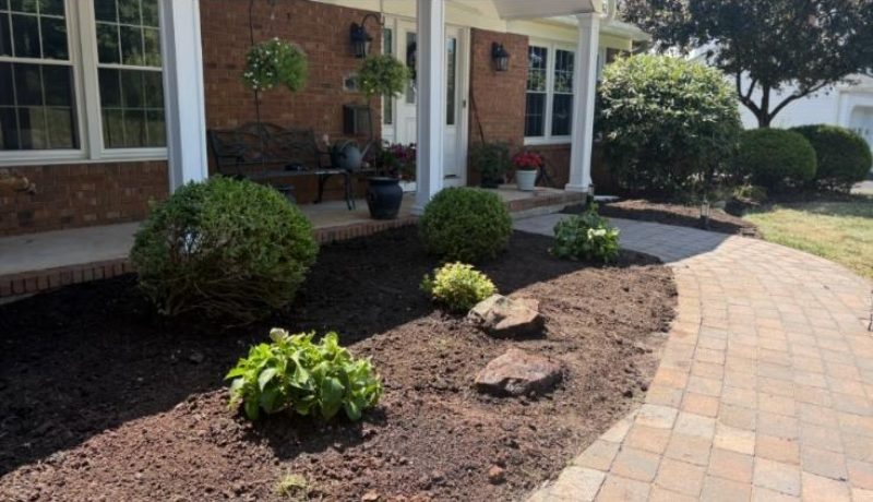 Freshly landscaped garden bed beside a brick house with newly planted shrubs, mulch, and a curved stone walkway leading to the porch.