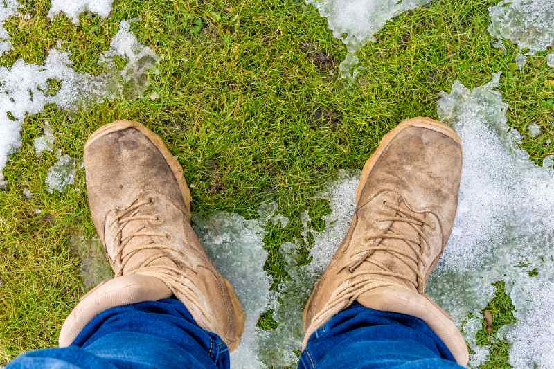 Worn tan boots standing on partially melted ice patches over soft green grass during winter thaw conditions.
