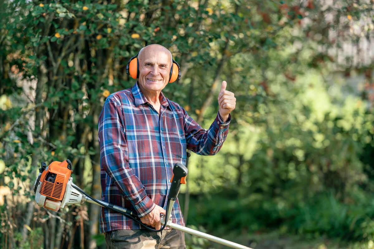 Elderly man wearing ear protection and holding grass trimmer giving thumb up with cheerful smile in lush garden setting.