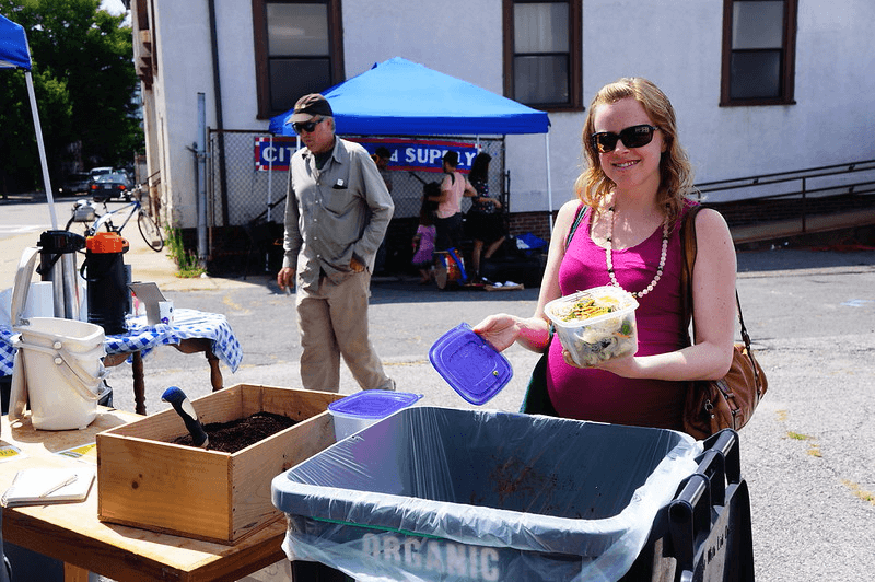 A woman dumps a Tupperware of food scraps in an organic recycling bin at a Boston farmers market