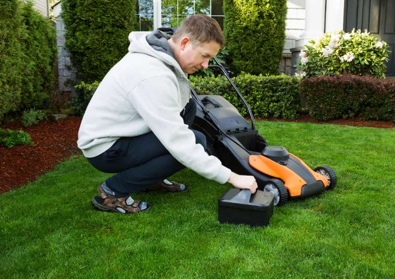 Man crouches on green lawn, inserting battery into orange electric lawn mower beside shrubs and house, preparing to mow today.