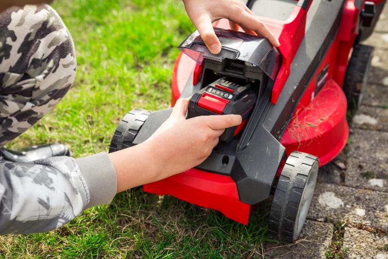 Close-up of hands inserting rechargeable battery into red electric lawn mower on grass beside paved path, preparing to mow.
