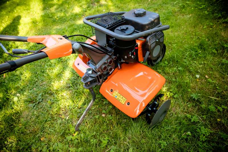 Gas powered garden tiller with orange housing resting on green grass, showing engine, handles, tines, and wheels ready for soil cultivation.