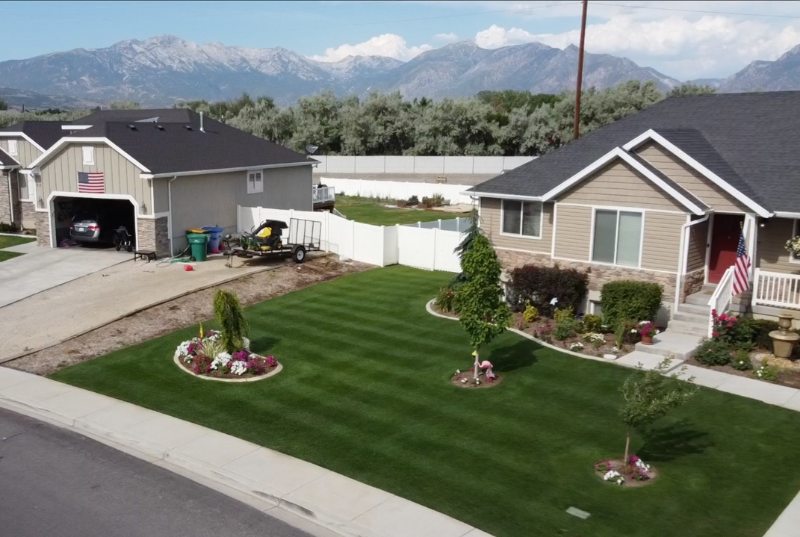 Aerial view of a suburban Utah home with a striped, golf course-like lawn, flower beds, neighboring houses, and mountains beyond.