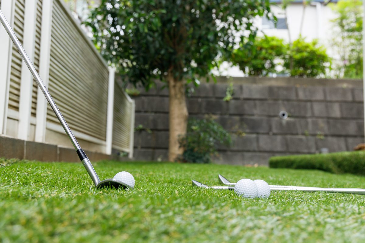 Golf club and balls rest on backyard grass beside fallen clubs, with fence, tree, and blurred garden wall in background.