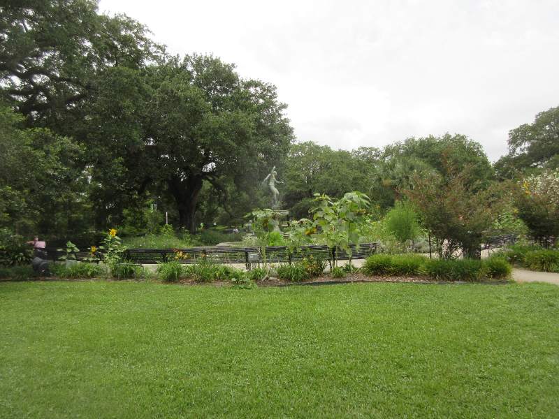 Wide green lawn bordered by garden beds, benches, and large trees, with a fountain statue rising in the background beneath gray sky.