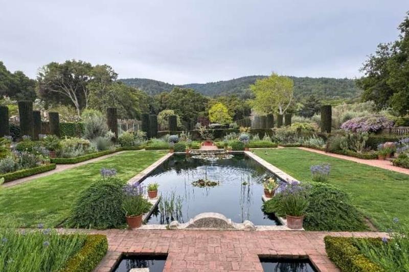 Wide view of Filoli Sunken Garden with reflecting pools, brick paths, clipped hedges, layered plantings, and wooded hills under gray sky.