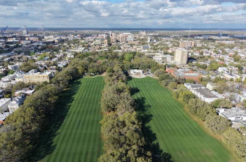 Aerial view of Forsyth Park with broad green lawns, tree-lined promenade, surrounding Savannah neighborhood, and cloudy sky stretching to the horizon.