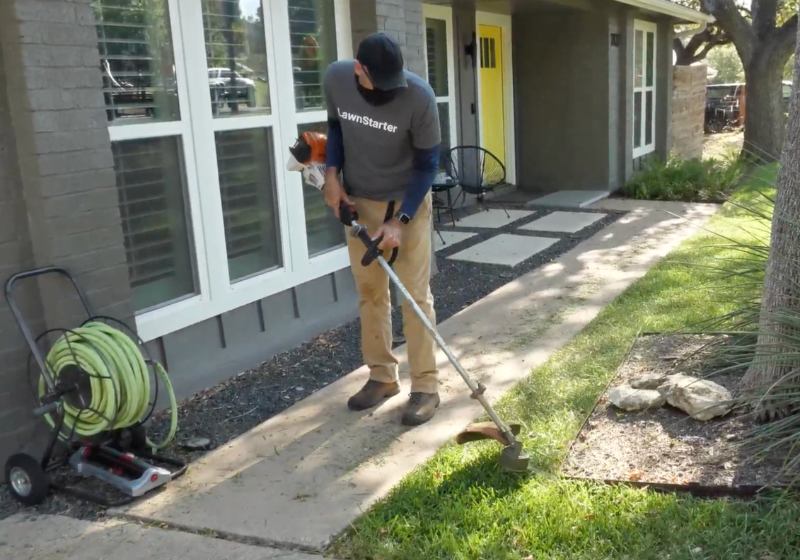 LawnStarter pro Justin Stultz uses a string trimmer to edge grass along a walkway beside a residential home.