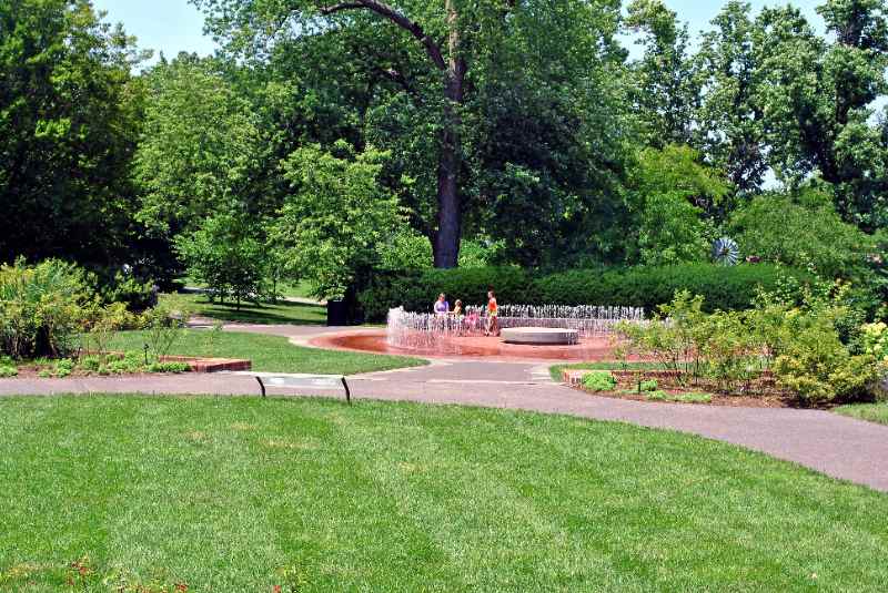 Missouri Botanical Garden lawn and winding paths frame a circular splash fountain, where children play amid trees and planted garden beds.