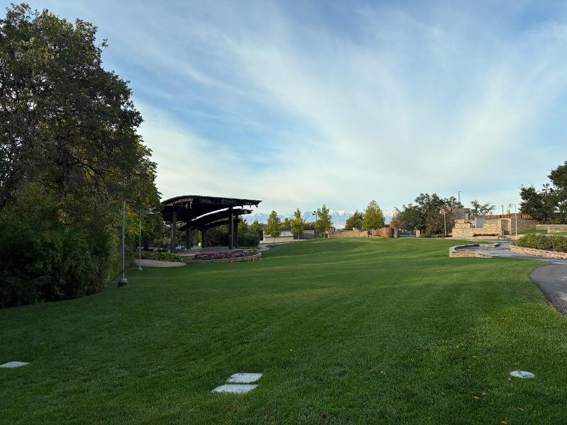 Morning view of Red Butte Garden and Arboretum lawn, pavilion, trees, pathway, and distant mountains beneath a streaked blue sky.