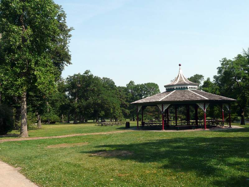 Tower Grove Park lawn with historic open pavilion, surrounding trees, picnic tables, and curving path beneath a clear bright summer sky.