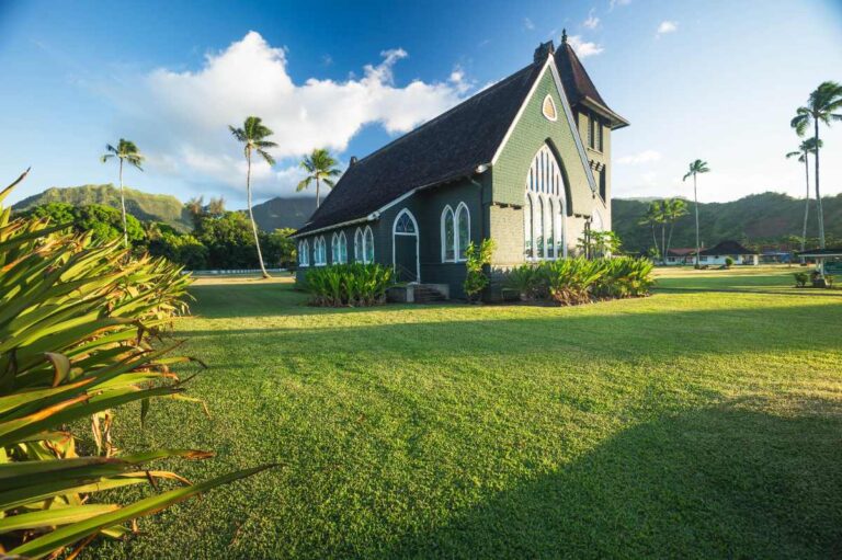 Green Waiʻoli Huiʻia Church in Hanalei, Kauai, Hawaii, with palm trees, mountain backdrop, lawn, and bright blue sky.