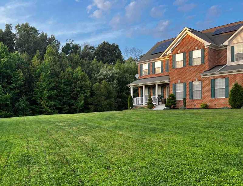 Large brick house beside a freshly mowed green lawn, with striped grass, porch, solar panels, and trees behind.