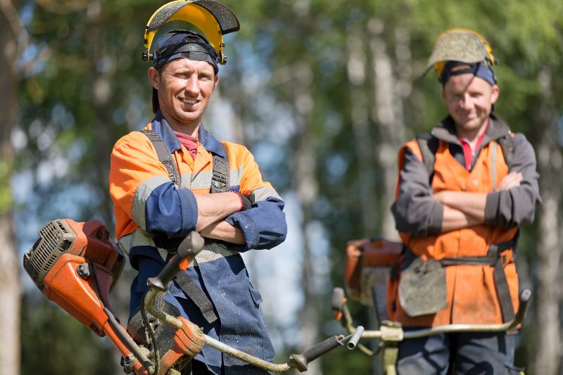 Two local lawn care workers stand with string trimmers, wearing orange safety vests and face shields in a wooded outdoor area.