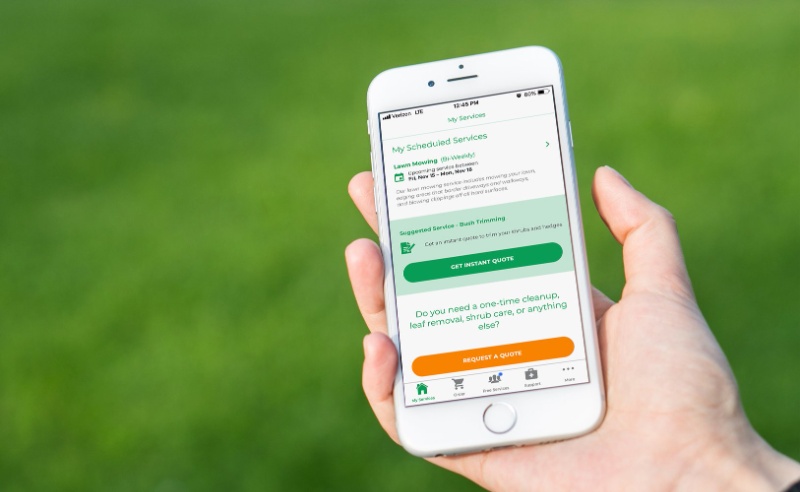 Hand holding smartphone showing lawn care service scheduling app against a green grass background, with quote request options visible.