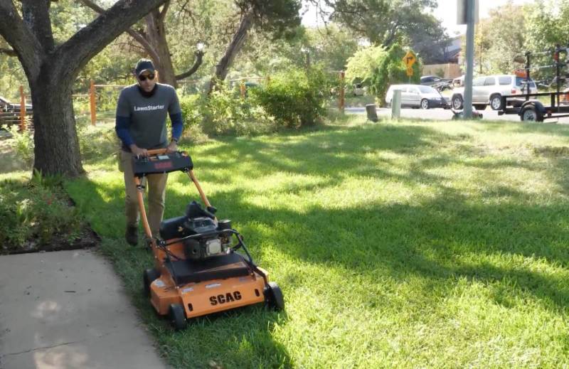 Justin Stultz pushes an orange mower along a sidewalk, cutting tall grass in a shaded residential yard.
