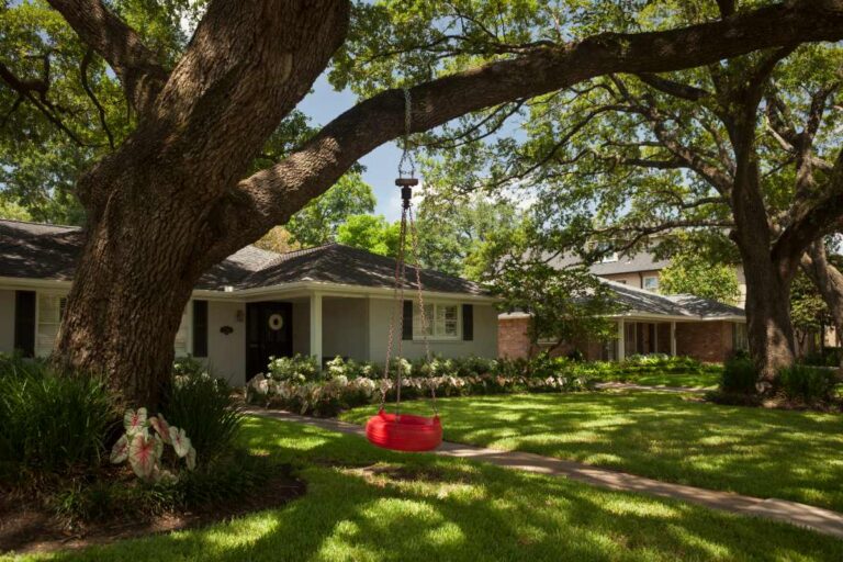 Houston home landscape with large shade trees, green lawn, garden beds, walkway, and a red swing hanging from an oak.