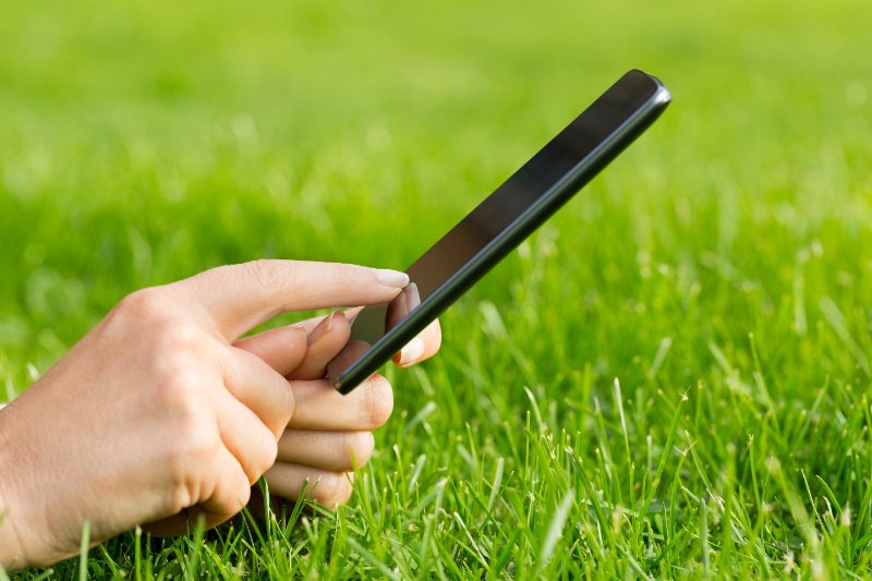 A close-up of a person's hand holding a black smartphone horizontally over a lush green grass lawn, with a finger tapping the screen.