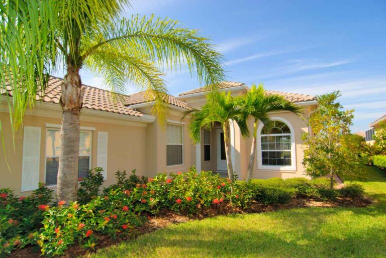 A single-story Florida home with a tan stucco exterior and terracotta tile roof, surrounded by tall palm trees, red flowering shrubs, and a well-manicured green lawn under a bright blue sky.