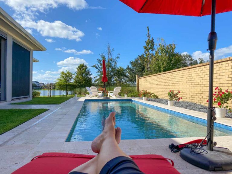 A first-person view of someone relaxing on a red lounge chair beside a backyard swimming pool, feet in the foreground, with white Adirondack chairs, red umbrellas, potted red flowers, a brick privacy wall, green lawn, and a lake visible beyond the iron fence under a blue sky.