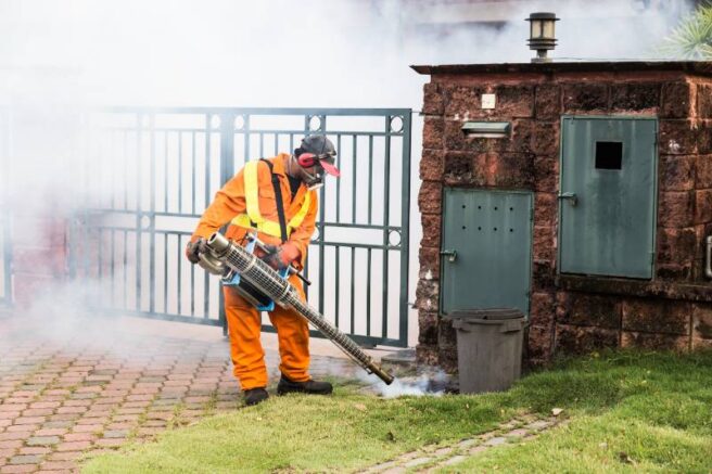A pest control worker in an orange jumpsuit, yellow safety vest, respirator, and ear protection using a fogging machine to spray insecticide along a residential lawn and driveway to kill mosquitoes.