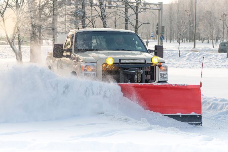 A tan pickup truck equipped with a bright red snow plow blade pushes a large wave of snow down a snow-covered road lined with bare winter trees, with yellow fog lights illuminated.