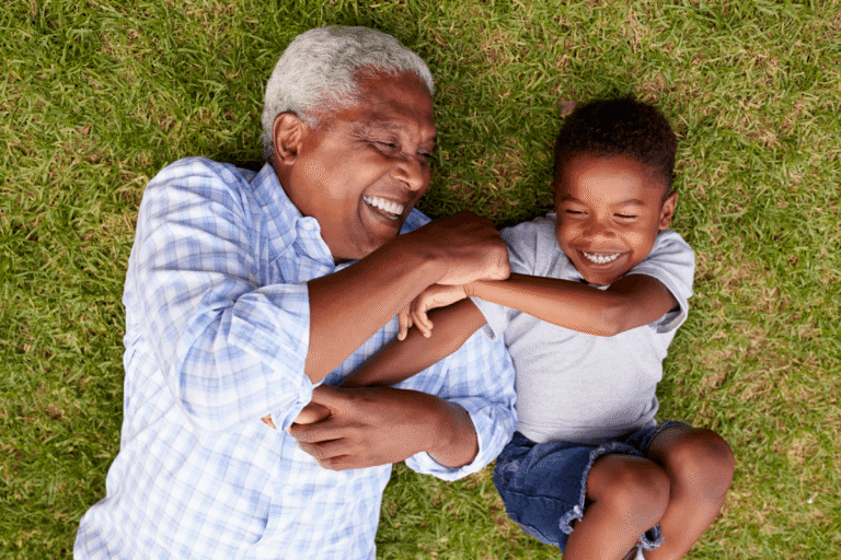 An overhead view of a smiling older man with white hair and a young boy lying together on the grass, laughing and embracing each other while playing outdoors.
