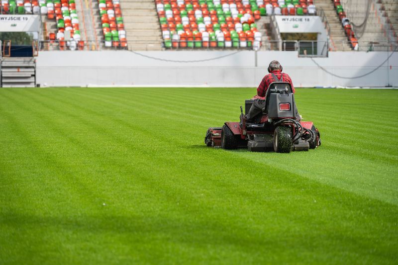 A groundskeeper in a red flannel shirt rides a commercial reel mower across a vast, perfectly striped green football stadium field, with rows of empty white, orange, and green stadium seats visible in the background.