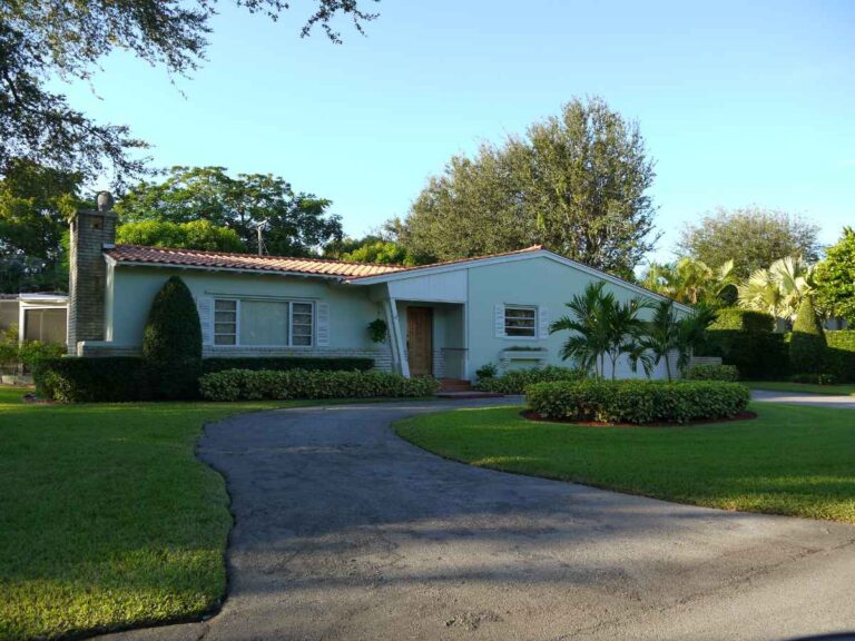 A mint green mid-century ranch home in Coral Gables, Miami, with a terracotta tile roof, brick chimney, curved asphalt driveway, manicured hedges, areca palms, red mulch beds, and a lush green lawn under a clear blue sky at golden hour.