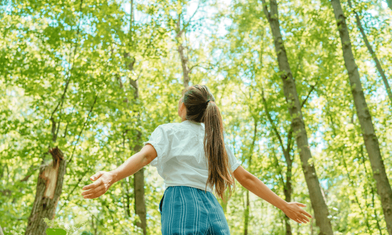 A woman with a long ponytail, wearing a white blouse and striped blue pants, stands with her back to the camera and arms outstretched among tall green trees in a sunlit forest.