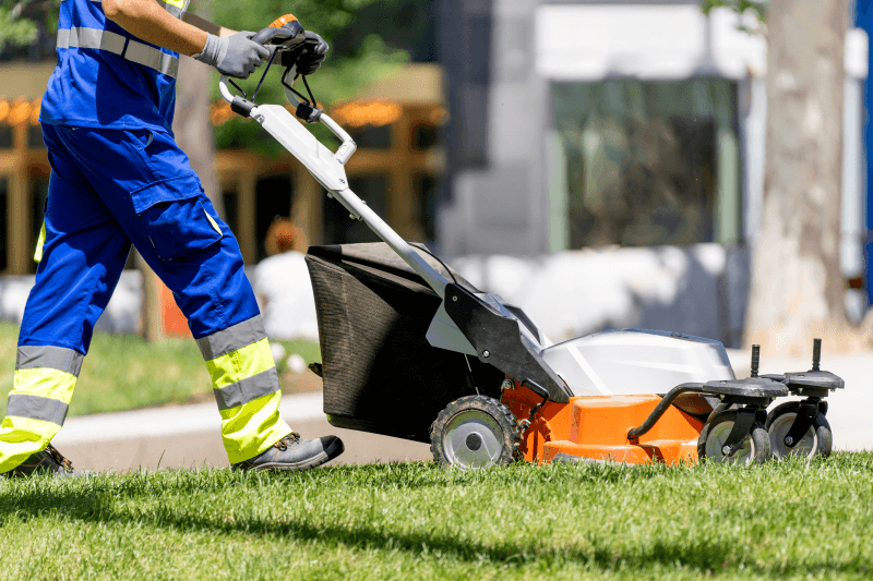 A worker wearing blue coveralls with high-visibility safety stripes pushes an orange and black lawn mower with a grass collection bag across a green lawn.