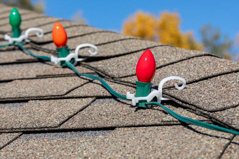 A close-up of colorful red, orange, and green Christmas string lights clipped to gray asphalt roof shingles with white plastic light clips.