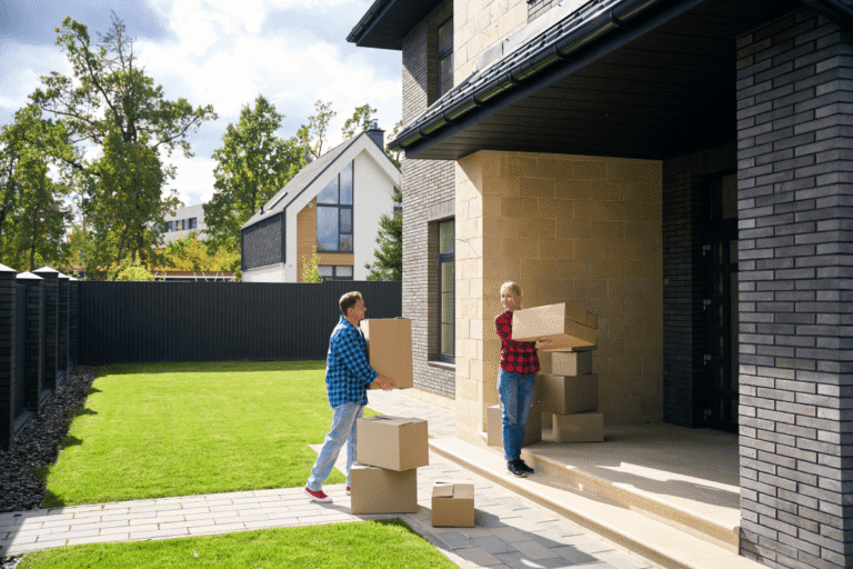 A couple carrying moving boxes toward the entrance of a modern two-story house with several more boxes stacked on the front steps and walkway.