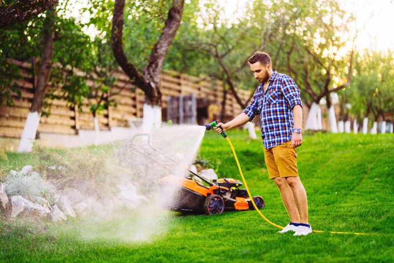 A man stands on a green lawn watering the grass with a hose and spray nozzle.