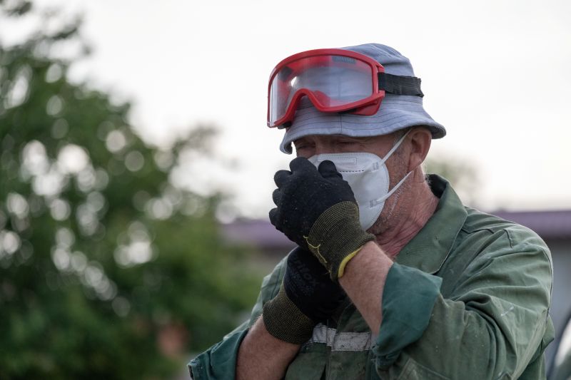 A man in a green work jacket wears a protective respirator mask, safety goggles pushed up on a bucket hat, and work gloves.