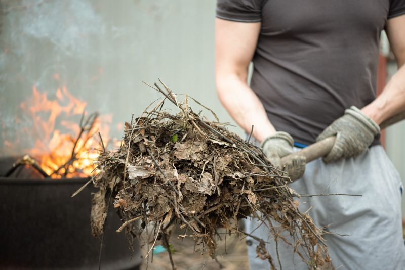 A person in work gloves holds a pitchfork piled with dry leaves, twigs, and garden debris beside a metal barrel with flames burning inside.